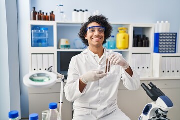 Young hispanic man wearing scientist uniform holding test tubes at laboratory