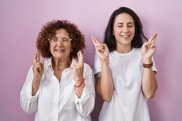 Hispanic mother and daughter wearing casual white t shirt over pink background gesturing finger crossed smiling with hope and eyes closed. luck and superstitious concept.