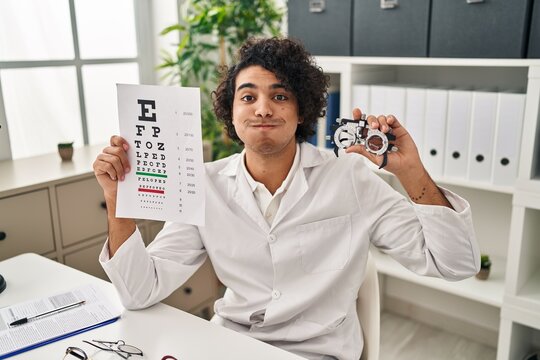 Hispanic Man With Curly Hair Holding Optometry Glasses And Eyesight Test Puffing Cheeks With Funny Face. Mouth Inflated With Air, Catching Air.