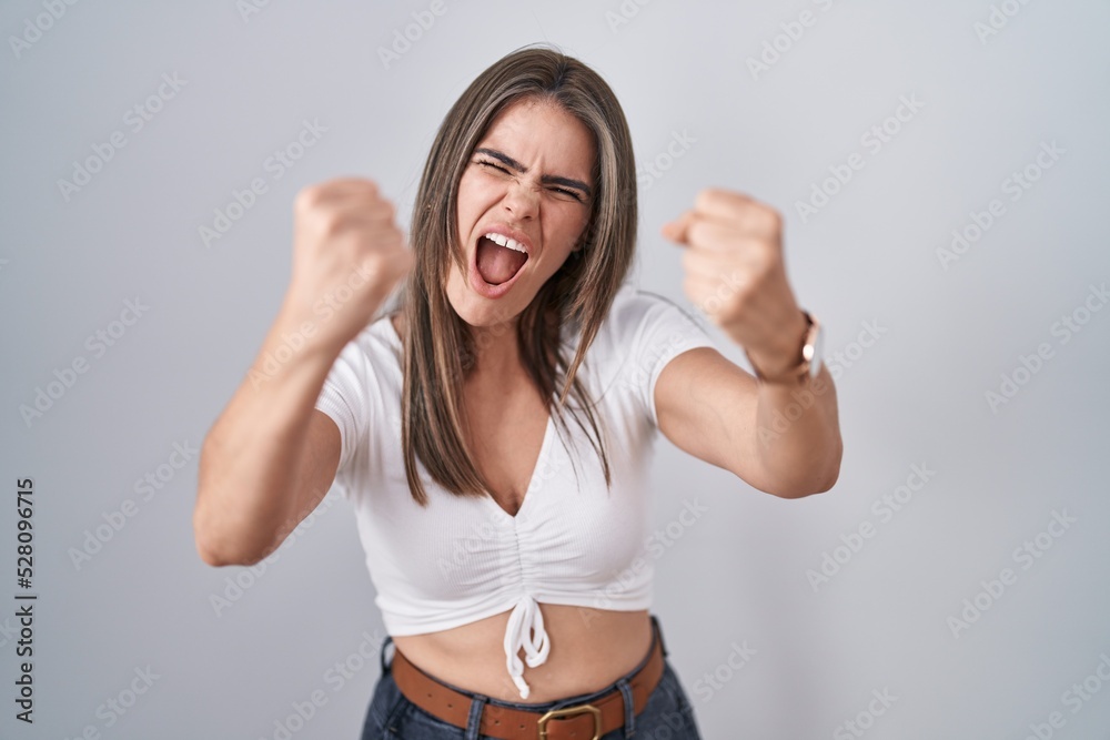 Wall mural Young beautiful woman wearing casual white t shirt angry and mad raising fists frustrated and furious while shouting with anger. rage and aggressive concept.