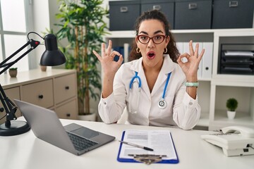 Young hispanic woman wearing doctor uniform and stethoscope looking surprised and shocked doing ok approval symbol with fingers. crazy expression