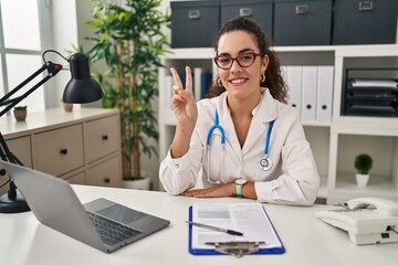 Young hispanic woman wearing doctor uniform and stethoscope smiling with happy face winking at the camera doing victory sign with fingers. number two.