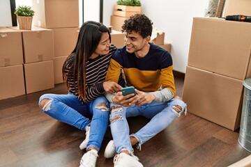 Young latin couple using smartphone sitting on the floor at new home.