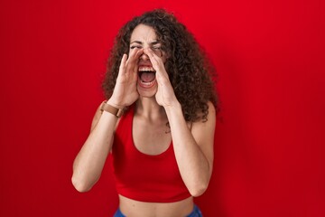 Hispanic woman with curly hair standing over red background shouting angry out loud with hands over...