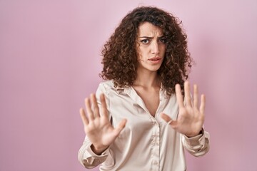 Hispanic woman with curly hair standing over pink background moving away hands palms showing refusal and denial with afraid and disgusting expression. stop and forbidden.