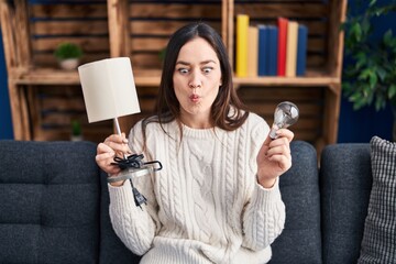 Young brunette woman holding led lightbulb and lamp making fish face with mouth and squinting eyes,...