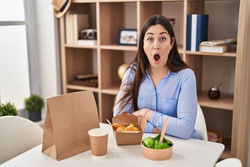 Young brunette woman eating take away food at home scared and amazed with open mouth for surprise, disbelief face
