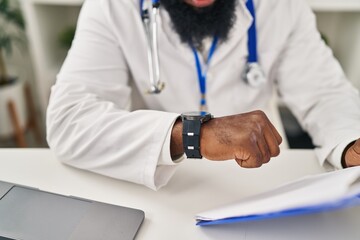 Young african american man wearing doctor uniform looking watch at clinic