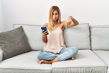 Obraz premium Blonde beautiful young woman sitting on the sofa at home using smartphone pointing down looking sad and upset, indicating direction with fingers, unhappy and depressed.