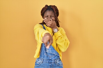 African woman standing over yellow background laughing at you, pointing finger to the camera with...