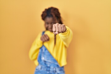African woman standing over yellow background punching fist to fight, aggressive and angry attack,...