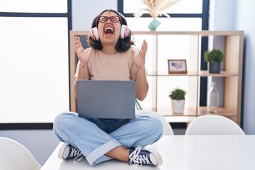Fototapeta na wymiar Young hispanic woman using laptop sitting on the table wearing headphones celebrating mad and crazy for success with arms raised and closed eyes screaming excited. winner concept
