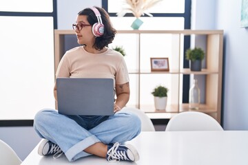 Fototapeta na wymiar Young hispanic woman using laptop sitting on the table wearing headphones looking to side, relax profile pose with natural face with confident smile.