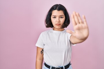 Young hispanic woman wearing casual white t shirt over pink background doing stop sing with palm of the hand. warning expression with negative and serious gesture on the face.