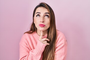 Young hispanic woman standing over pink background thinking concentrated about doubt with finger on chin and looking up wondering