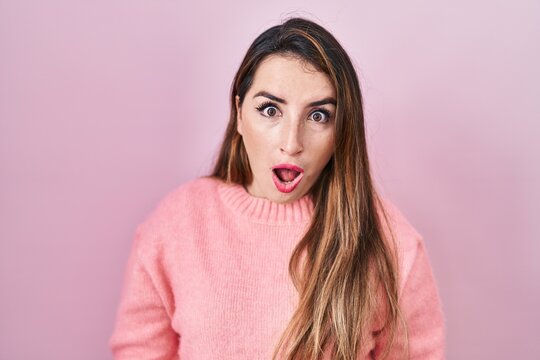 Young hispanic woman standing over pink background in shock face, looking skeptical and sarcastic, surprised with open mouth
