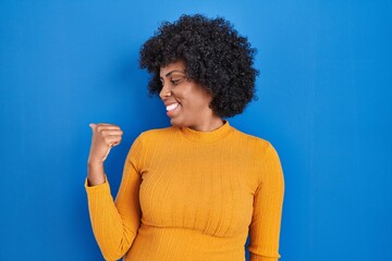 Black woman with curly hair standing over blue background smiling with happy face looking and pointing to the side with thumb up.