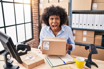 Black woman with curly hair working at small business ecommerce holding box angry and mad screaming...