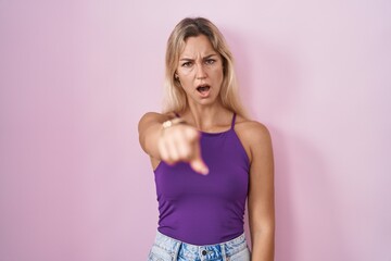Young blonde woman standing over pink background pointing displeased and frustrated to the camera,...