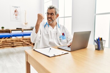 Middle age hispanic therapist man working at pain recovery clinic with laptop annoyed and frustrated shouting with anger, yelling crazy with anger and hand raised