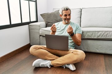 Middle age hispanic man using laptop sitting on the floor at the living room doing money gesture...
