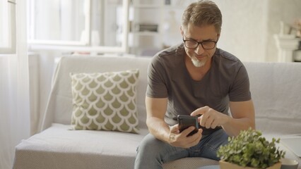 Older man at home, sitting on couch in living room, using phone, checking social media. Portrait of happy, mature age, middle age, mid adult caucasian man in 50s, smiling.
