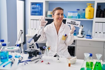 Middle age hispanic woman working at eco friendly laboratory doing ok sign with fingers, smiling friendly gesturing excellent symbol