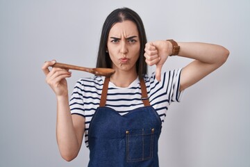 Young brunette woman wearing apron tasting food holding wooden spoon with angry face, negative sign...