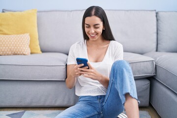 Young hispanic woman using smartphone sitting on floor at home