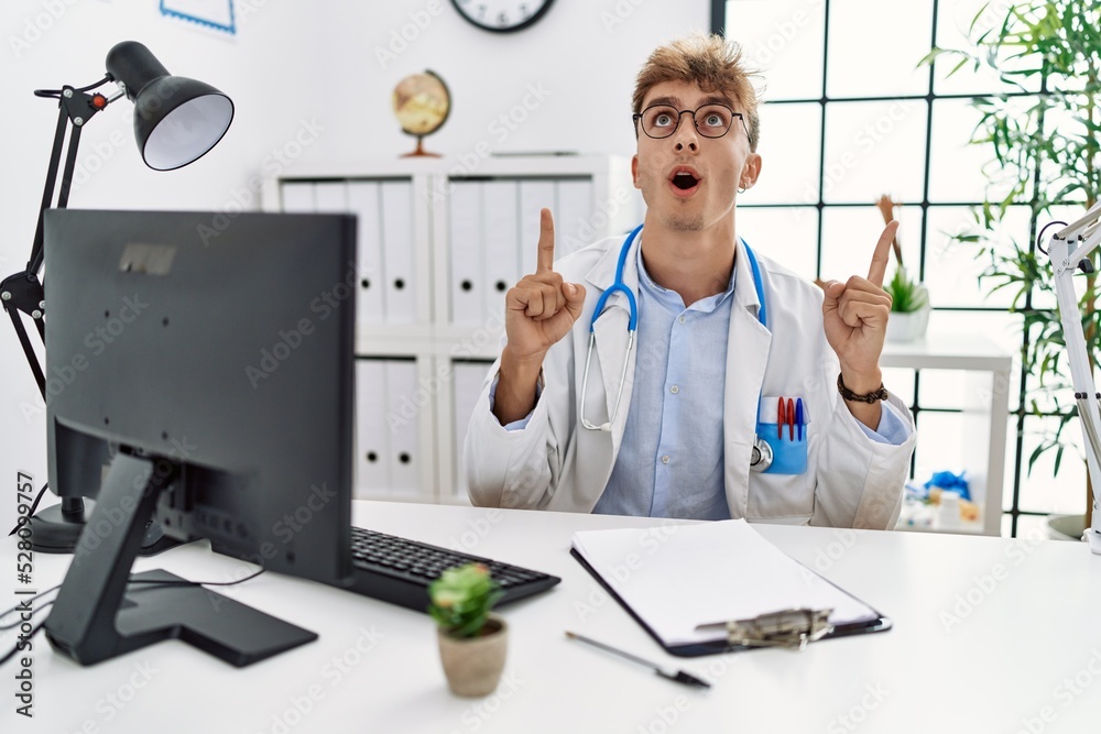 Canvas Prints Young caucasian doctor man working at the clinic amazed and surprised looking up and pointing with fingers and raised arms.