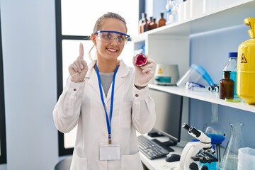 Young woman working at scientist laboratory holding geode smiling with an idea or question pointing finger with happy face, number one