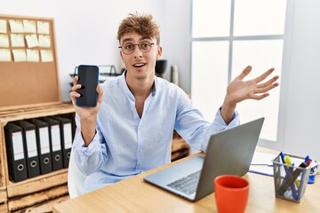 Young caucasian businessman working using laptop and showing smartphone celebrating achievement with happy smile and winner expression with raised hand