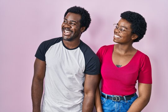 Young african american couple standing over pink background looking away to side with smile on face, natural expression. laughing confident.