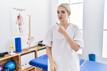 Young caucasian woman working at pain recovery clinic pointing aside worried and nervous with forefinger, concerned and surprised expression