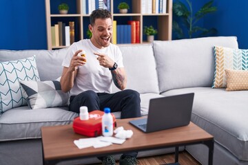 Young hispanic man watching video on laptop on how to use first aid kit smiling happy pointing with...