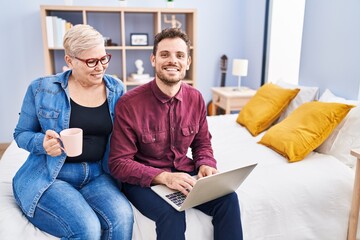 Mother and son using laptop drinking coffee sitting on bed at bedroom