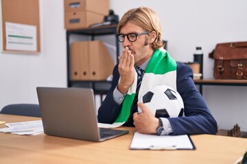 Caucasian man with mustache working at the office supporting football team covering mouth with...
