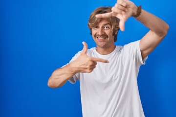 Middle age man standing over blue background smiling making frame with hands and fingers with happy face. creativity and photography concept.