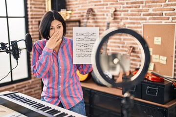 Young asian woman doing online music tutorial showing music sheet covering mouth with hand, shocked and afraid for mistake. surprised expression