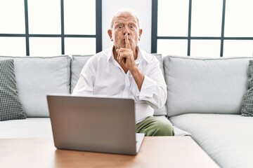 Senior man using laptop at home sitting on the sofa asking to be quiet with finger on lips. silence and secret concept.