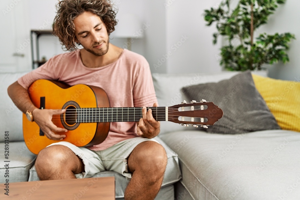 Canvas Prints Young hispanic man smiling happy playing classical guitar sitting on the sofa at home.