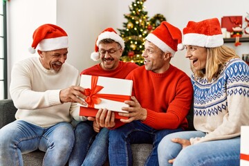 Fototapeta premium Group of middle age friends holding gifts sitting on the sofa by christmas tree at home.