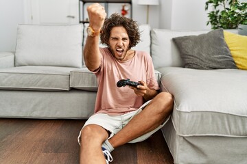 Young hispanic man playing video game holding controller at home annoyed and frustrated shouting...