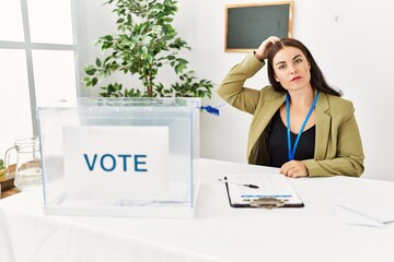 Young brunette woman sitting at election table with voting ballot confuse and wonder about...