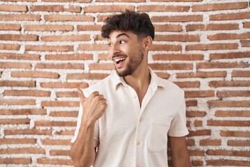 Arab man with beard standing over bricks wall background smiling with happy face looking and pointing to the side with thumb up.