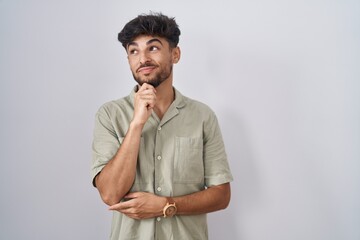 Arab man with beard standing over white background with hand on chin thinking about question, pensive expression. smiling with thoughtful face. doubt concept.