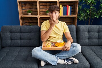 Arab man with beard sitting on the sofa at home eating breakfast covering mouth with hand, shocked...