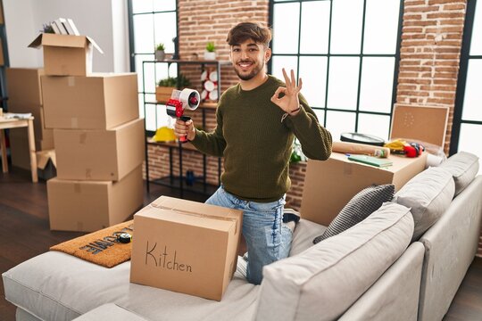 Arab man with beard moving to a new home closing cardboard box doing ok sign with fingers, smiling friendly gesturing excellent symbol