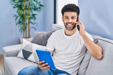 Young arab man talking on the smartphone using touchpad sitting on sofa at home