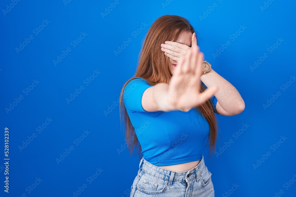 Canvas Prints redhead woman standing over blue background covering eyes with hands and doing stop gesture with sad